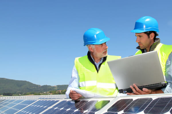 two men standing behind a pv module and talking with each other, one of them holding a laptop in his hands